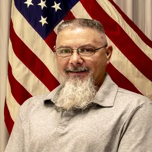 A man with long beard and glasses standing in front of an american flag.